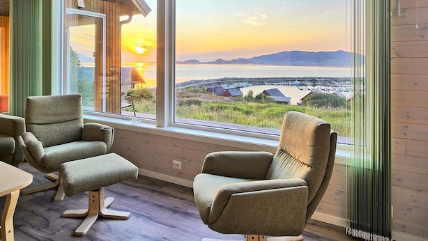 Ein Zimmer mit großem Fenster und Blick auf eine malerische Wasserlandschaft und ferne Berge. Zwei Polstersessel und ein Holztisch sind vorhanden.