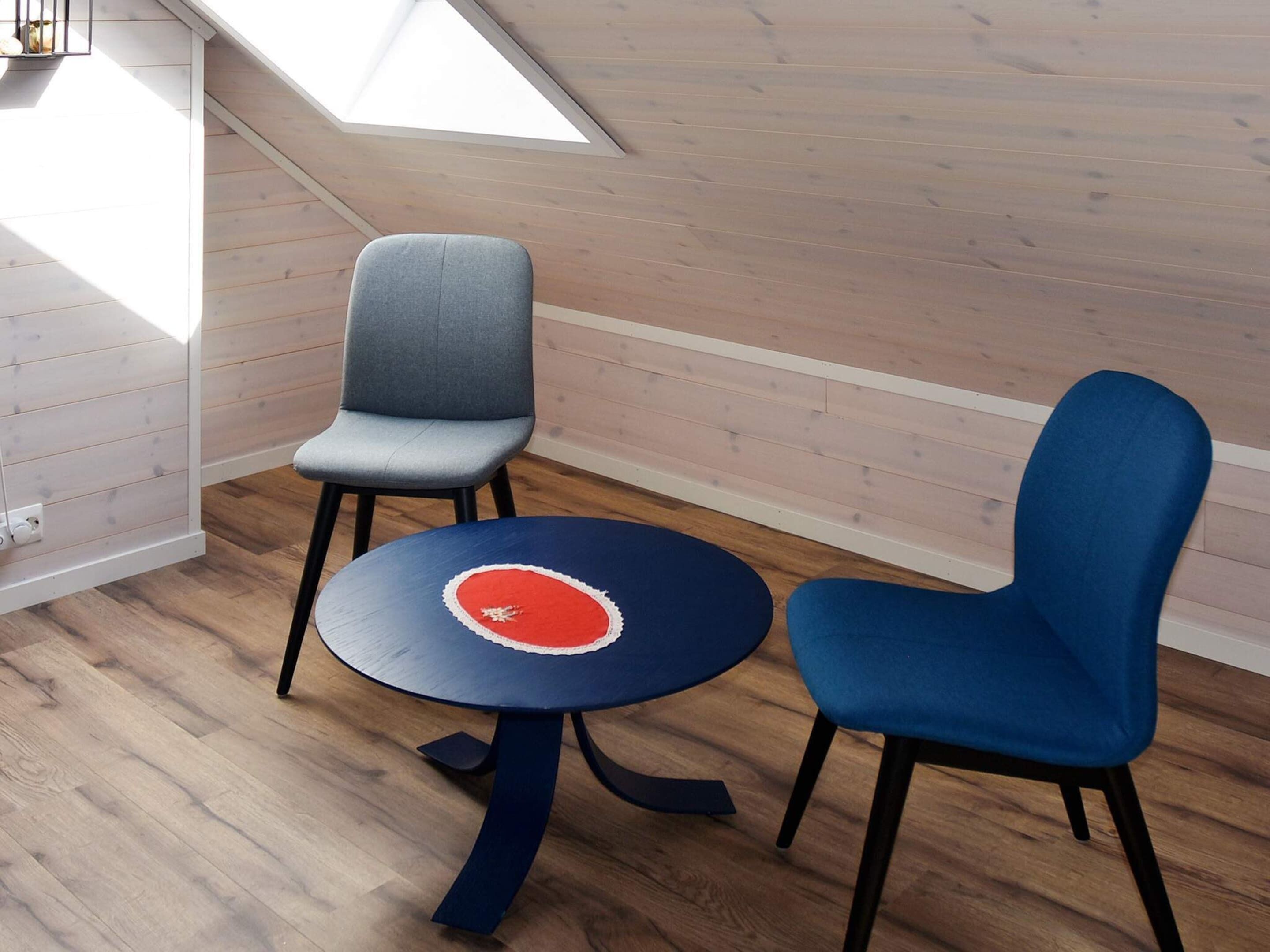 A room with a sloped ceiling, wooden walls, a round table with a red and white design, and two chairs.