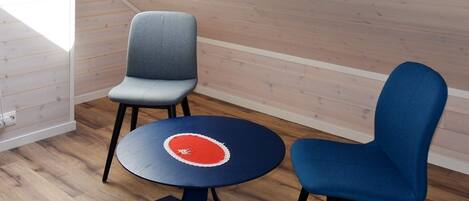 A room with a sloped ceiling, wooden walls, a round table with a red and white design, and two chairs.