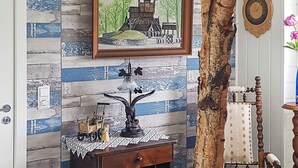 Una habitación con pared de madera rústica, un cuadro enmarco de una construcción tradicional, una mesita con una lámpara y una silla antigua.
