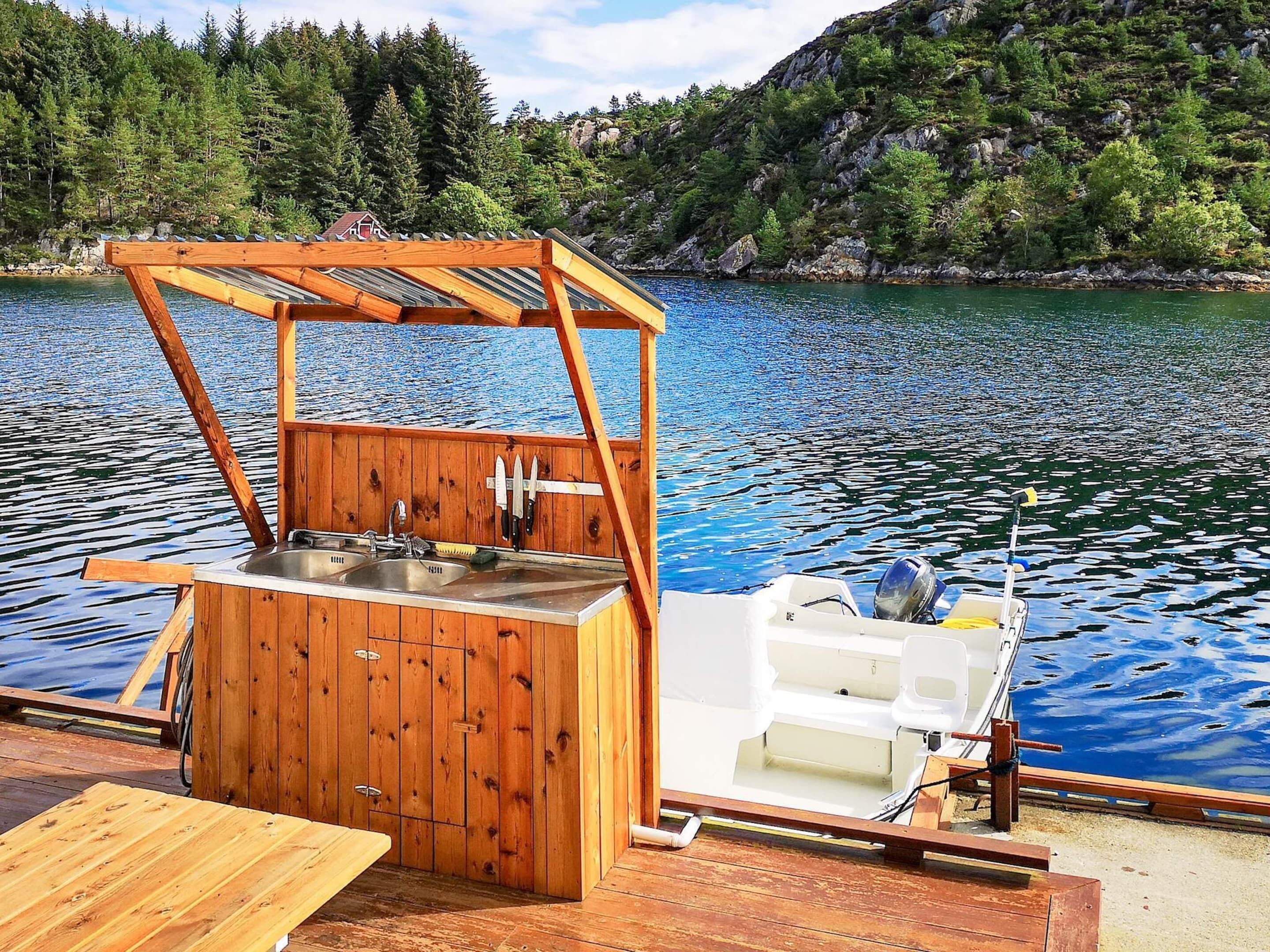 A wooden boat with a built-in kitchen area, including a sink and a small refrigerator, docked at a calm lake with forested hills in the background.