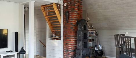 A spacious living room with a high ceiling, a brick fireplace, a wooden staircase, and a television on a white console.