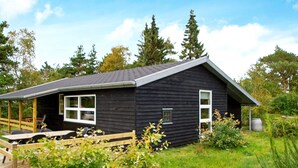 A small, dark wooden cabin with a single window and a door, surrounded by lush greenery and trees. - 6 Person Holiday Home in Skjern (Skjern)