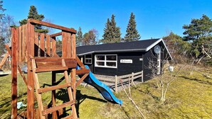 A wooden play structure with a slide in a grassy area, a black building with a sign reading "Fjelløser", and trees in the background. - 6 Person Holiday Home in Skjern (Skjern)