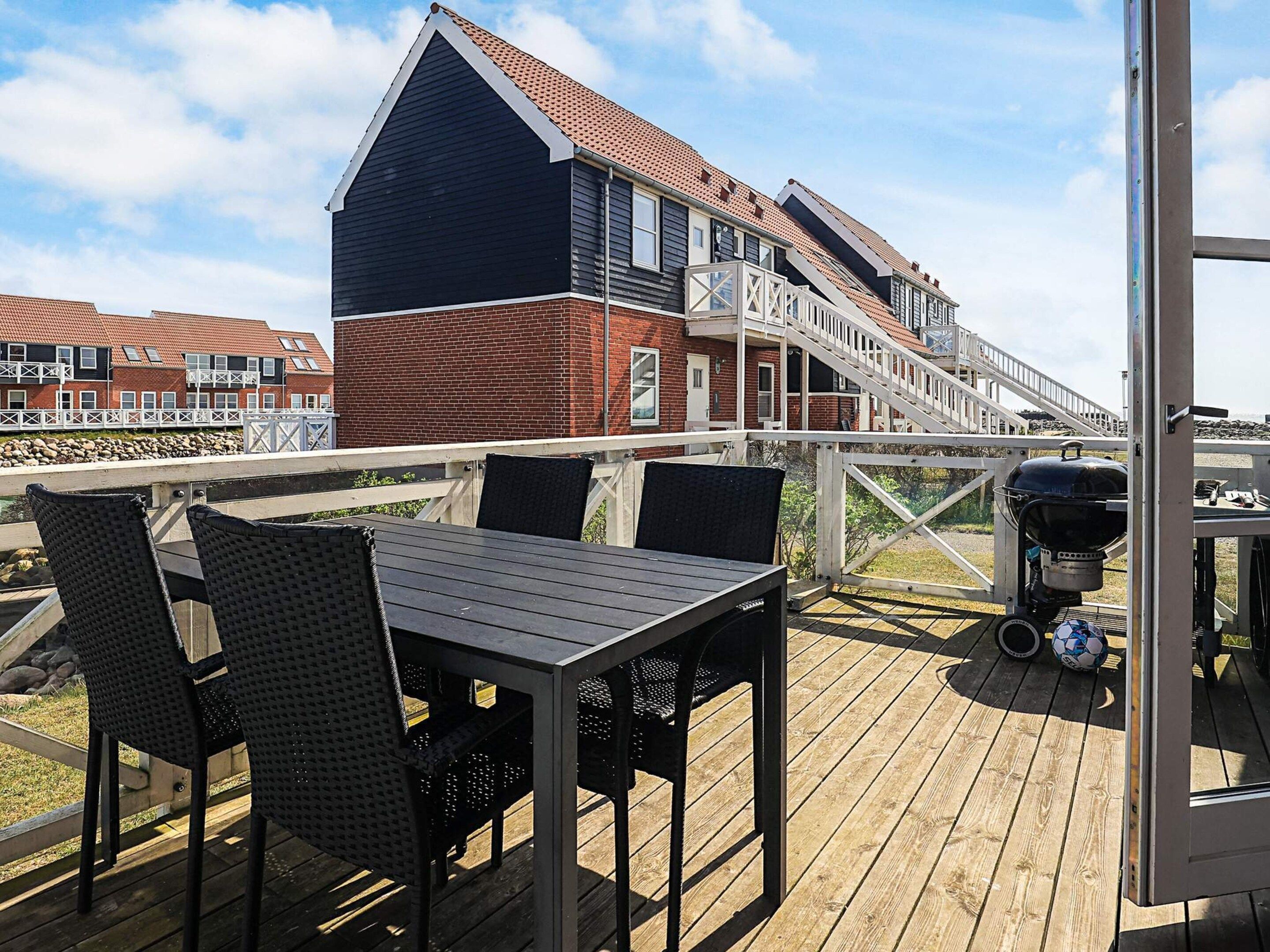 Une terrasse en bois avec des meubles d’extérieur, un barbecue et une vue sur un bâtiment en briques doté de nombreuses fenêtres et d’un toit en pente.