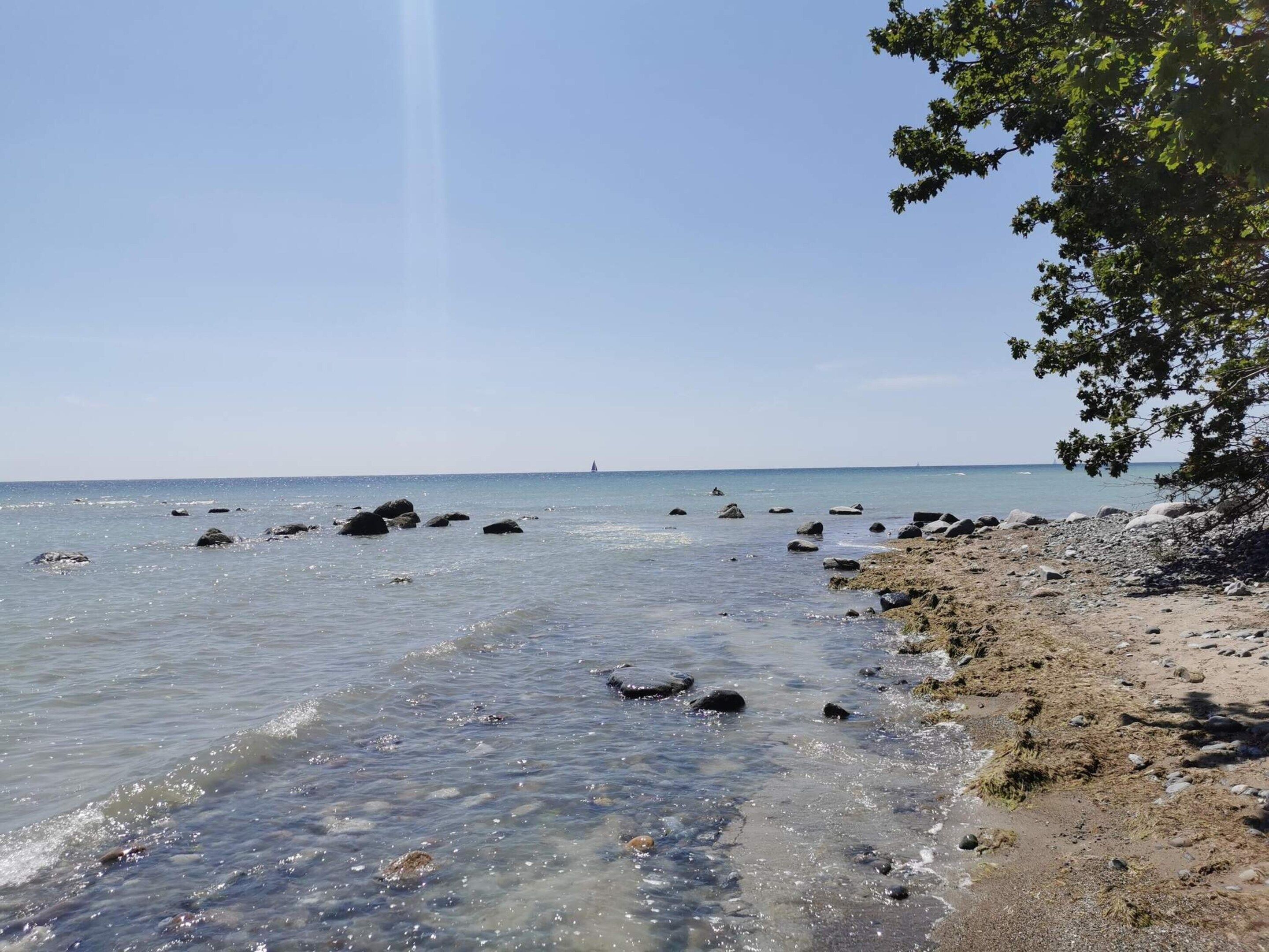 Une plage avec une eau claire, des rochers et un voilier qui se trouve au loin.