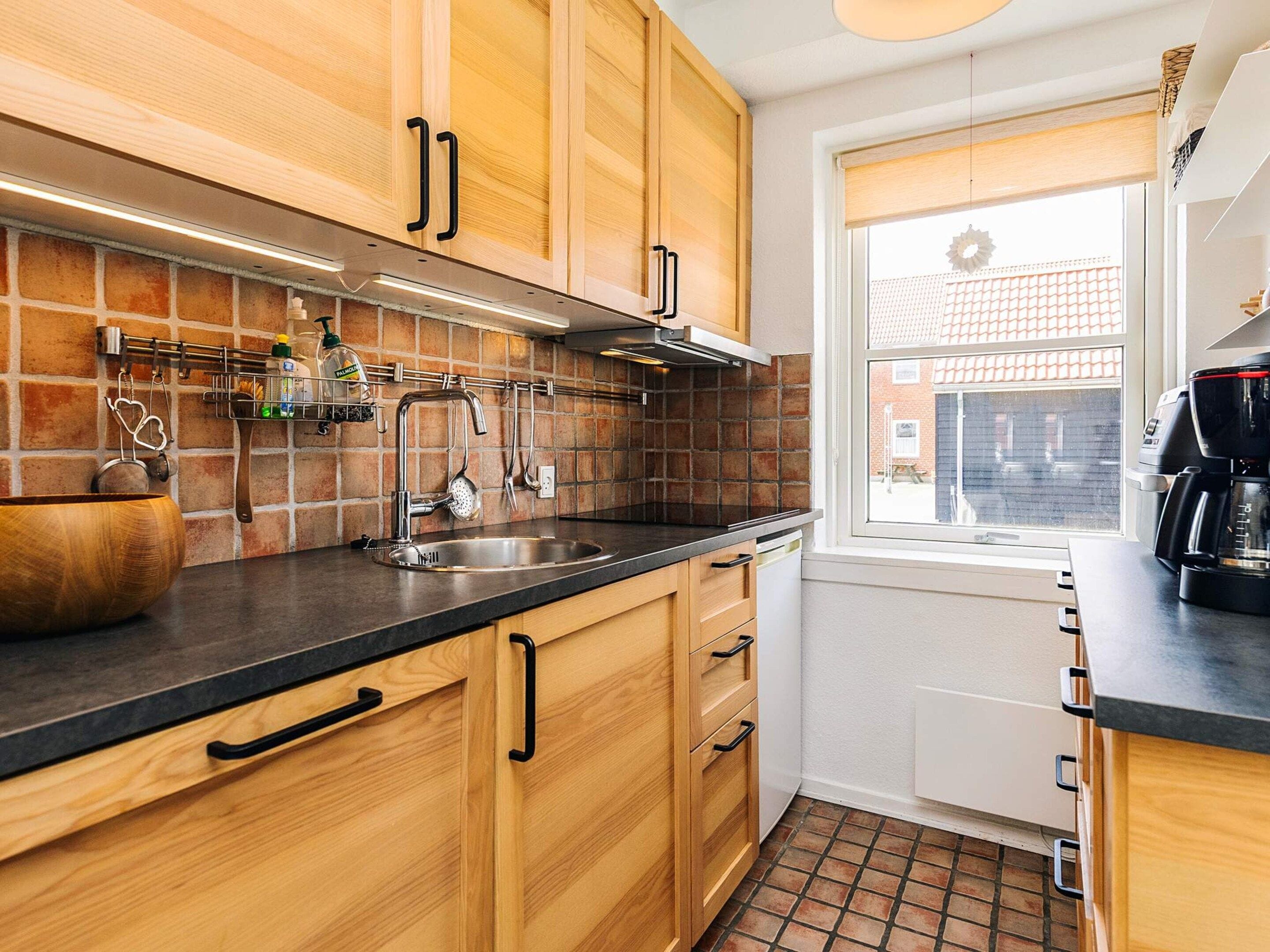 Een keuken met houten kastjes, een roestvrijstalen spoelbak en een raam met uitzicht op gebouwen.