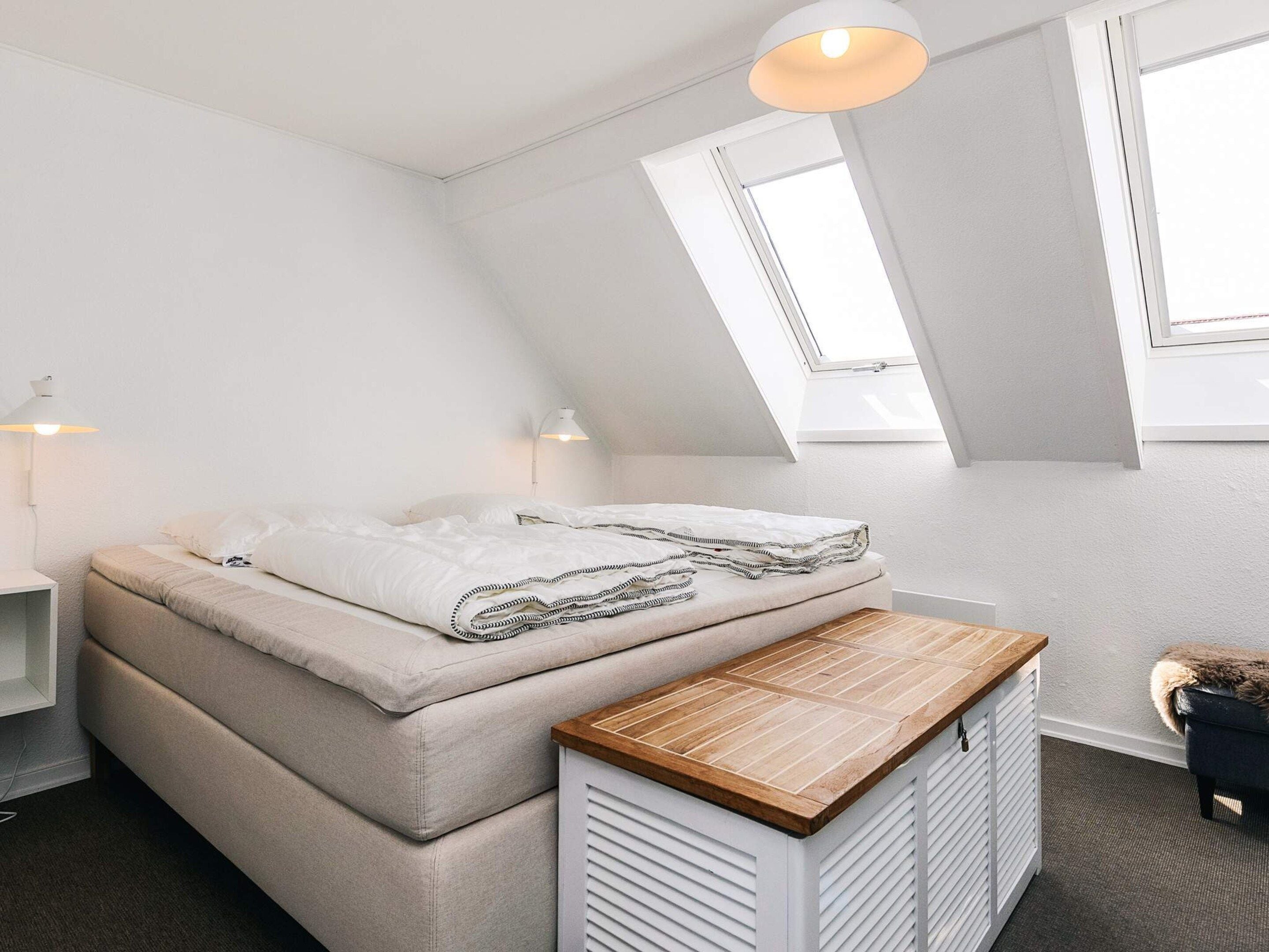 Une chambre située sous les toits, avec un plafond en pente, un lit doté d’un matelas beige et de draps blancs, un banc en bois au pied du lit et une lucarne.