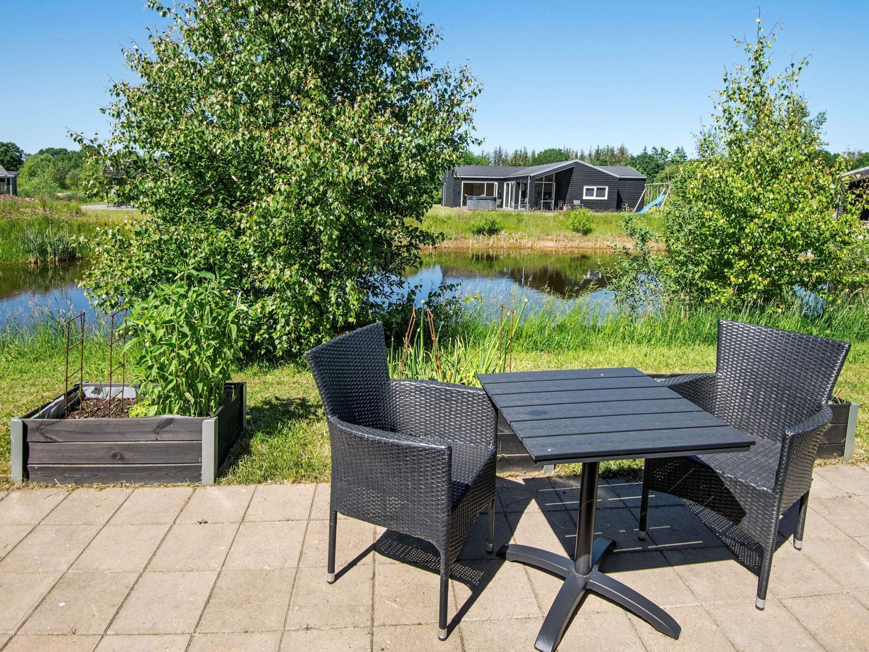 An outdoor patio with a table and two chairs, a raised planter box, and a view of a house across a body of water.