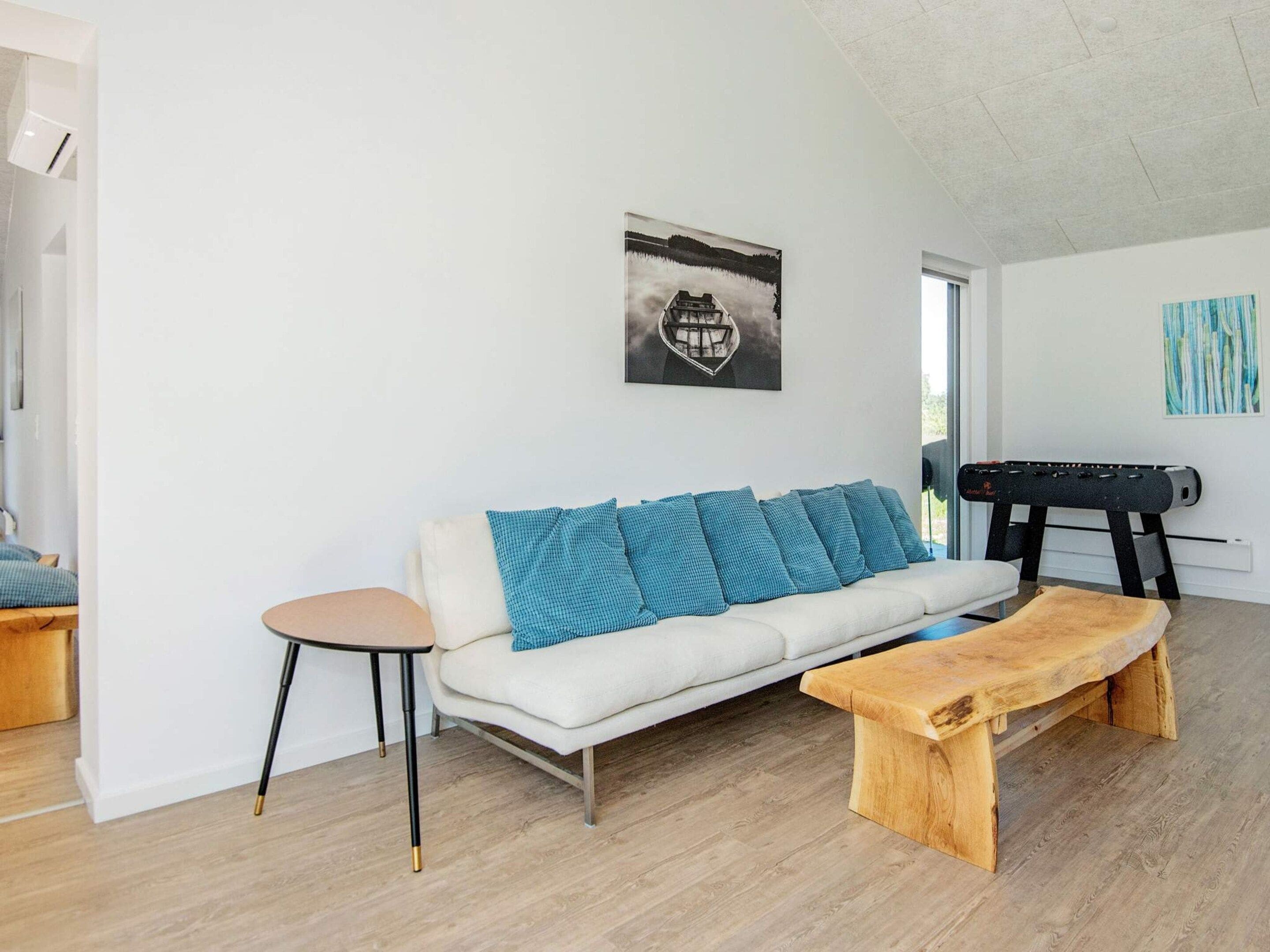Ein modernes Wohnzimmer mit einem weißen Sofa, blauen Kissen, einem runden Beistelltisch, einer Holzbank und einem Kicker.