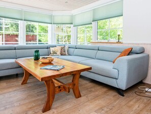 A room with a light blue sofa, a wooden table with a bowl and a magazine, and large windows with a view of greenery. - Vibrant Holiday Home in Tarm near Sea (Hemmet)