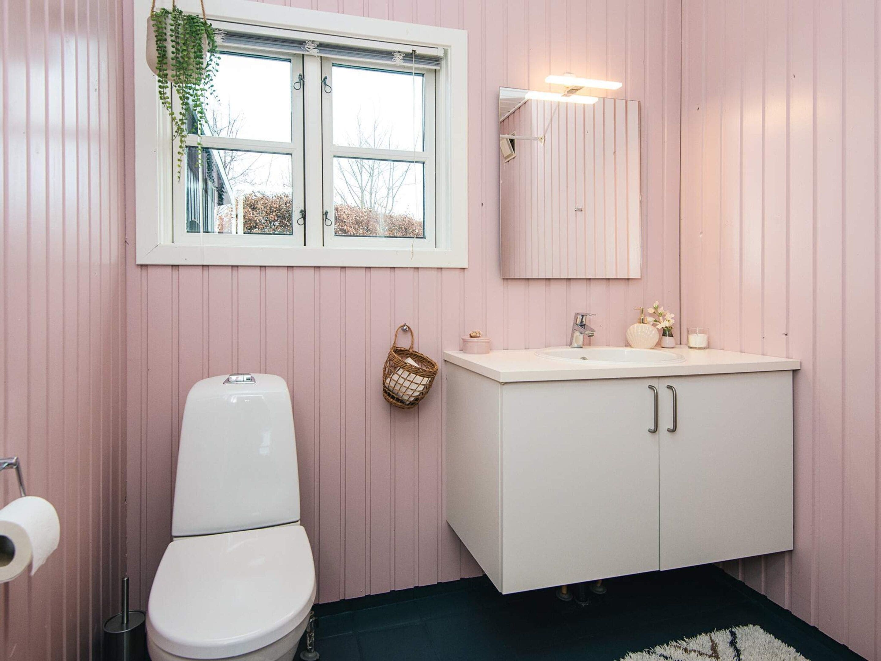 Un baño con paredes rosas, un inodoro blanco, un lavabo blanco con un espejo encima y una ventana con una planta colgando.