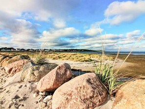 A beach with large rocks, sparse vegetation, and a distant shoreline with buildings. - Vibrant Holiday Home in Jutland near Sea (Glesborg)