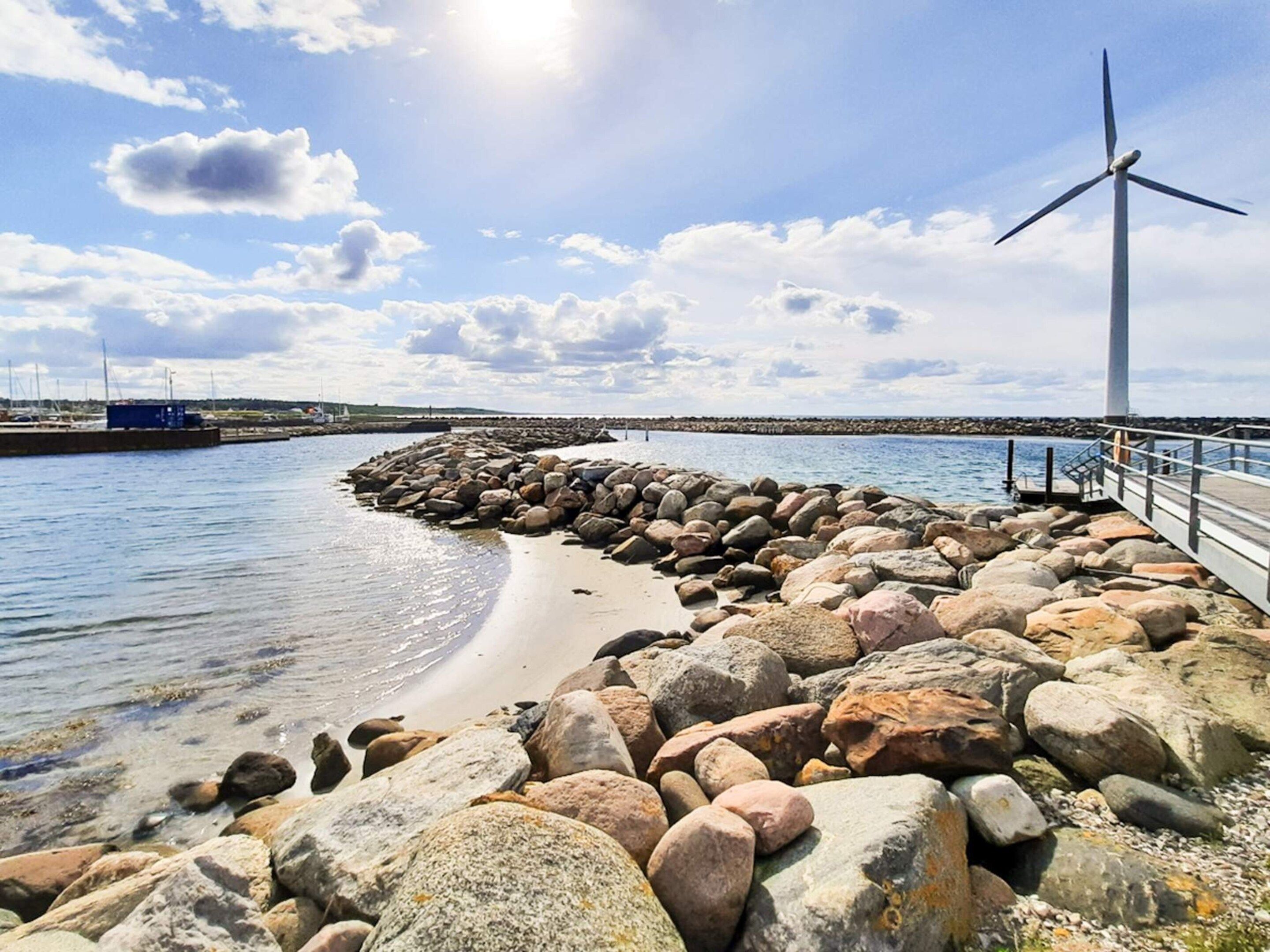 A coastal scene with a rocky breakwater, a wind turbine, and calm waters.