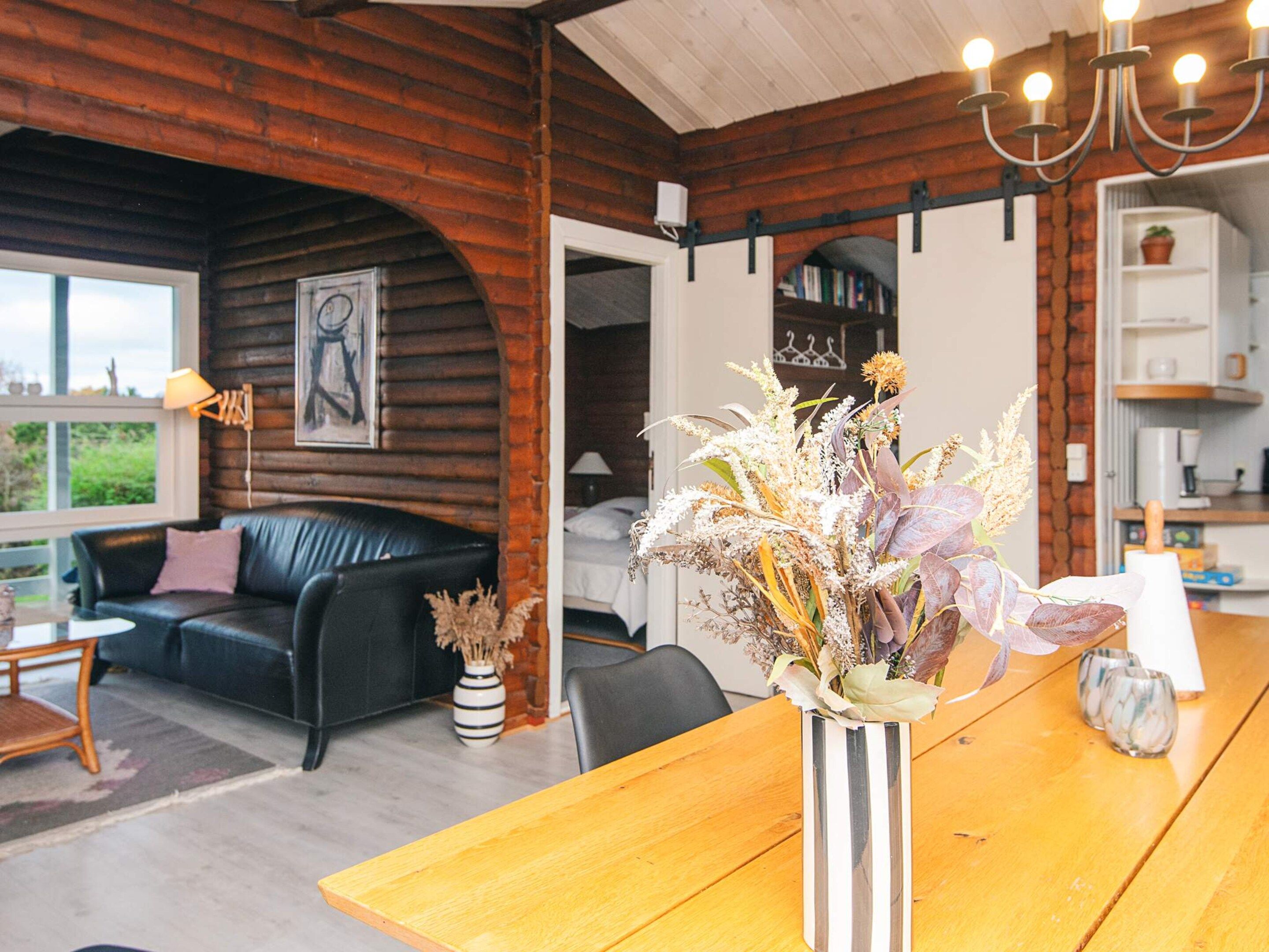 Una sala de estar acogedora con paredes de madera, un sofá de cuero negro, una mesa de comedor de madera y un jarrón con flores secas.
