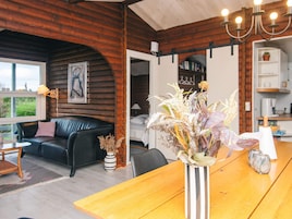 A cozy living area with a wooden interior, a black leather sofa, a wooden dining table, and a vase with dried flowers.