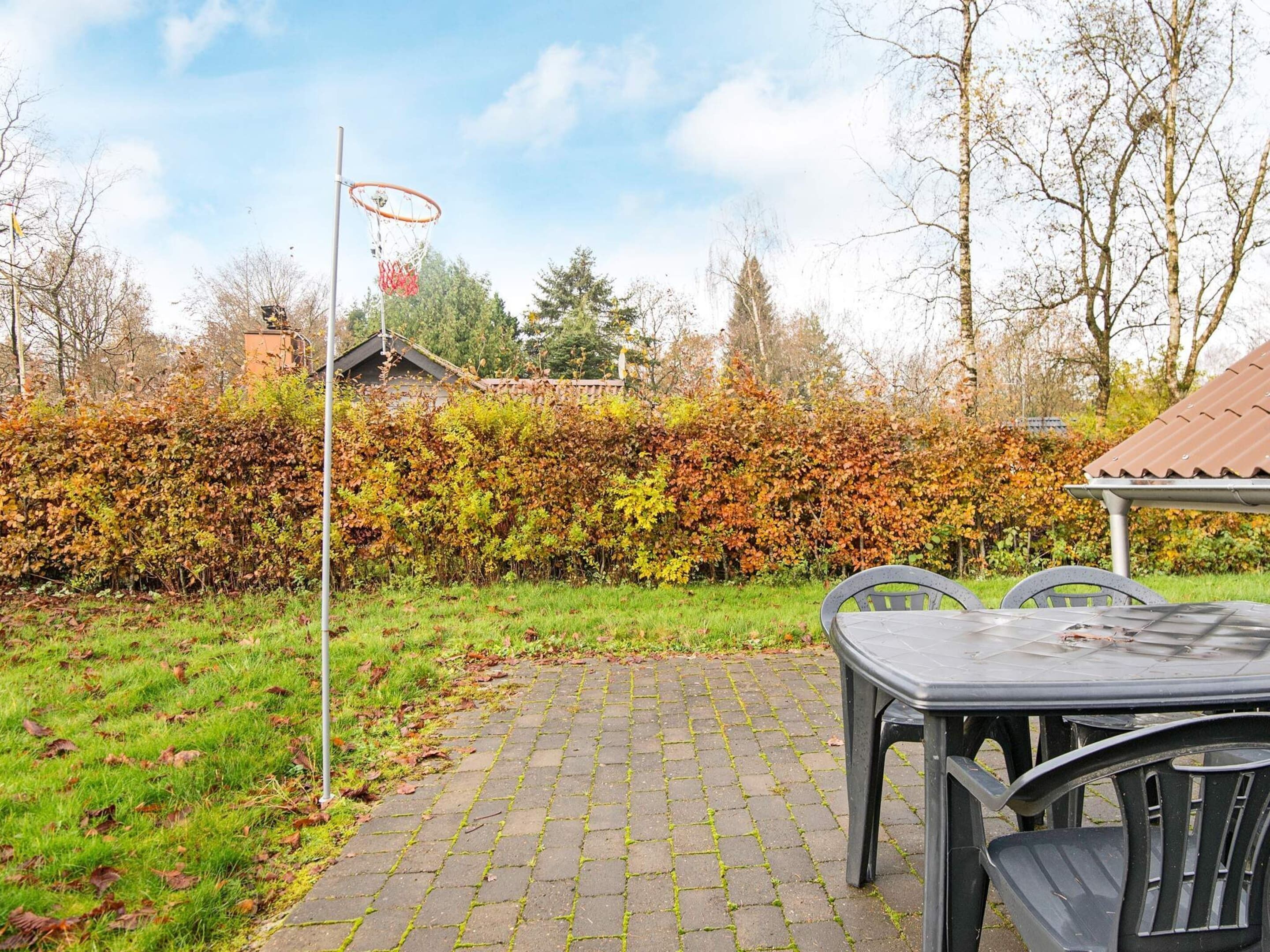 Une terrasse dallée avec une table et des chaises, un panier de basketball et une haie en arrière-plan.