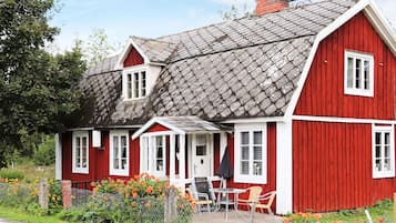 A red wooden house with a grey roof, a porch with chairs, and a fenced garden.