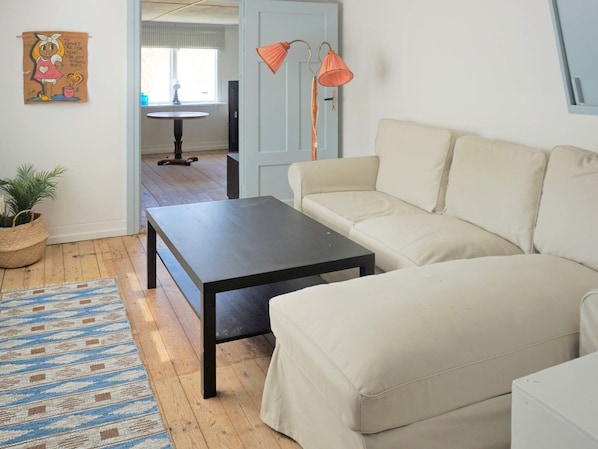 Un salon avec un canapé beige, une table basse en bois foncé, un tapis à motifs, une plante en pot et une lampe fixée au mur.