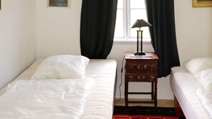 Quarto com duas camas de solteiro, mesa de cabeceira com abajur e janela com cortinas pretas.