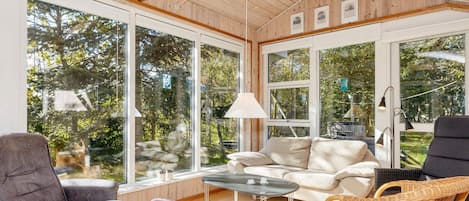 A sunlit room with a sofa, chairs, a table, and large windows offering a view of trees.