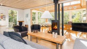 A cozy living room with a wooden ceiling, a fireplace, a TV, and a dining area with natural light from large windows. - 8 Person Holiday Home in Thyholm (Thyholm)