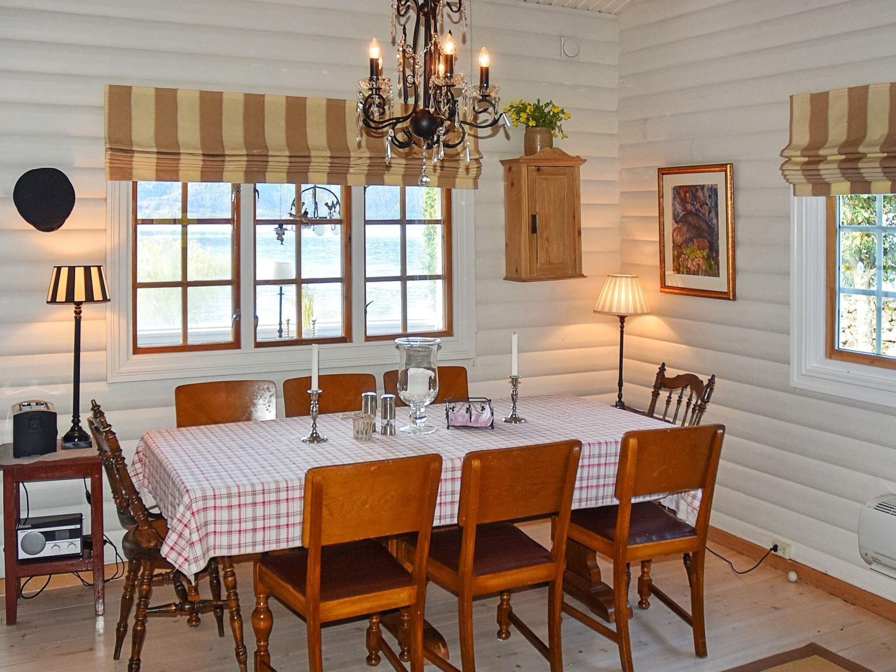 Un comedor con una mesa dispuesta para cuatro personas, rodeada de sillas de madera. Hay dos ventanas con cortinas, una araña y un aparador con una planta.