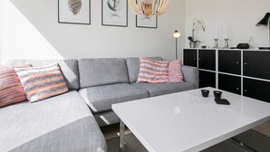 A modern living room with a grey sectional sofa, a white coffee table, and a black cabinet. There are two framed artworks on the wall, a pendant light, and a floor lamp.