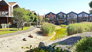 A row of modern wooden cabins with balconies, a gravel path, and a small pond.