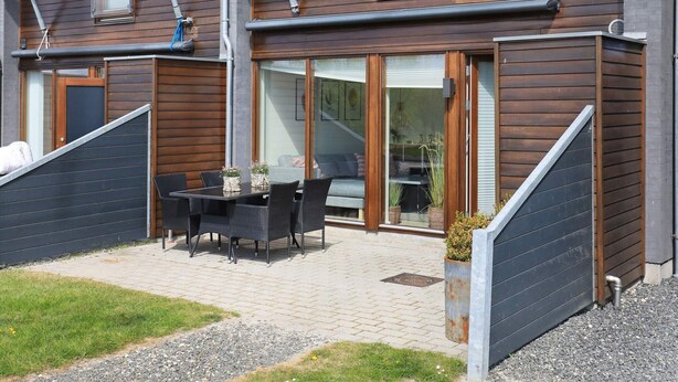 A modern two-story house with a patio area featuring a table and chairs, a sliding glass door entrance, and a small garden with potted plants.