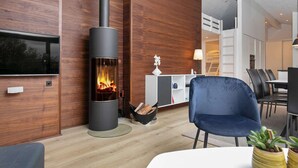 A modern living room with a wood-paneled wall, a black wood-burning stove, a flat-screen TV, a blue chair, and a dining area with a table and chairs. - 5 Star Holiday Home in Hjorring (Hjørring)