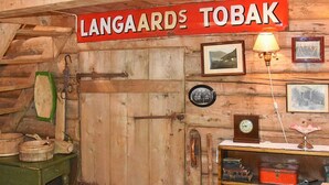 A rustic wooden interior with a sign reading 'LANGAARDS TOBAK,' a wooden door, a small table with a lamp, and a bookshelf with various items. - 3 Person Holiday Home in Bofjorden (Surnadal)