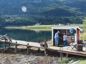 Um píer de madeira à beira do lago com bancos, uma pequena construção de madeira com churrasqueira, e uma casa ao fundo.