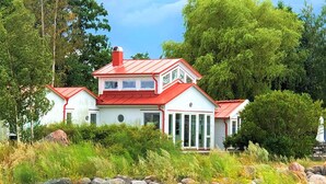 Une maison blanche avec un toit rouge, entourée de verdure et bordée d’une côte rocheuse.