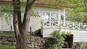 Ein weißes Gebäude mit einer Stein-Fundamentmauer, einer Holzterrasse mit Geländer und einem Baum im Vordergrund.