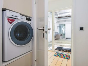A laundry room with a front-loading washer and dryer, a door leading to a balcony with a colorful mat, and a glimpse of a modern interior with a chair and a glass door. - 5 Star Holiday Home in Ebeltoft (Ebeltoft)