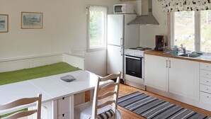Et kompakt køkken med et hvidt køleskab, komfur og mikroovn. Der er et spisebord med stole, et vindue med gardiner og en tæppe med et mønster på gulvet.