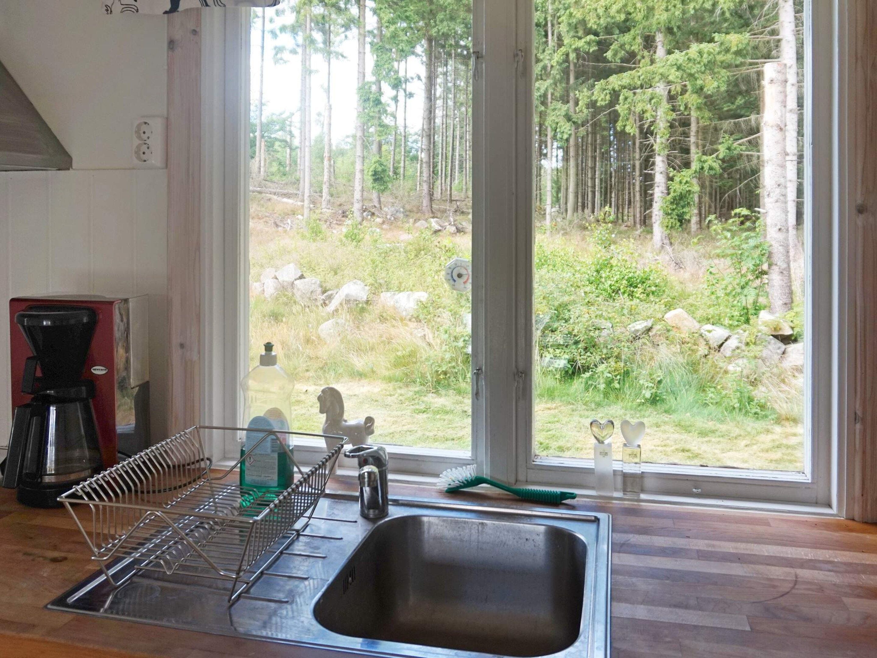 Une cuisine équipée d’un évier en acier inoxydable, d’un égouttoir à vaisselle, d’une machine à café et offrant une vue sur une forêt par la fenêtre.