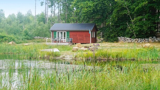 En lille, rød hytte med veranda, omgivet af en sø og frodig vegetation.