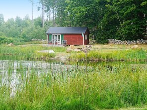 A small red cabin with a porch, surrounded by a pond and dense greenery.