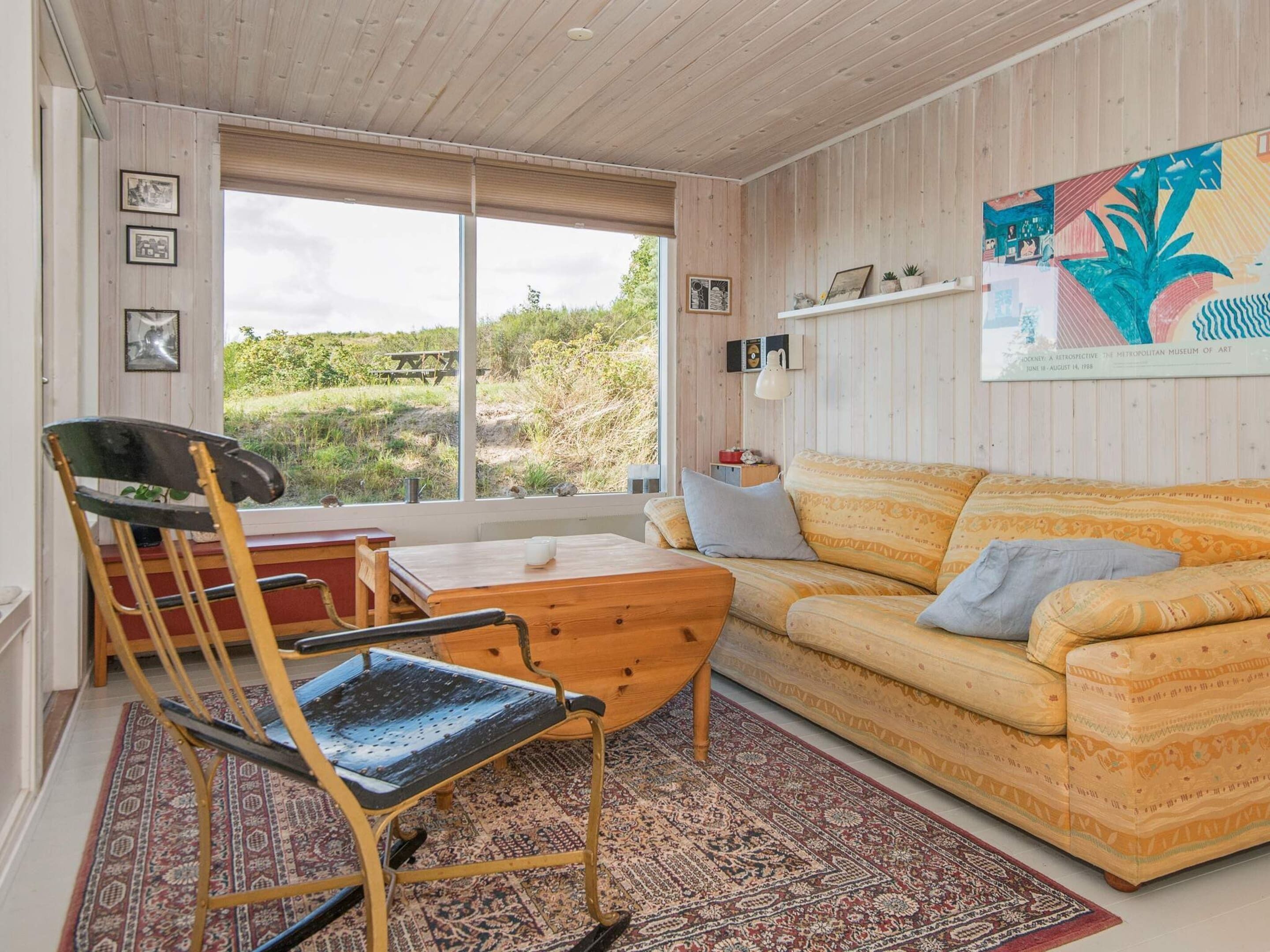 Un salon chaleureux avec un plafond en bois, un canapé à motifs, un fauteuil à bascule et une grande fenêtre donnant sur un paysage verdoyant.