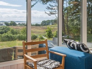 A room with a view of a grassy field and trees, a wooden chair, a blue sofa with patterned cushions, and large windows.