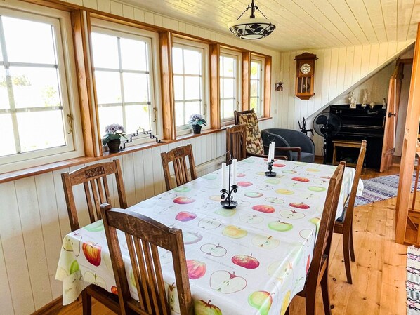 A dining area with a table covered in a fruit pattern tablecloth, wooden chairs, a clock on the wall, and a piano in the background.