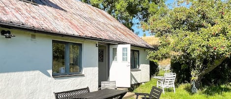 A white cottage with a metal roof, a wooden table, and two benches in the front yard.
