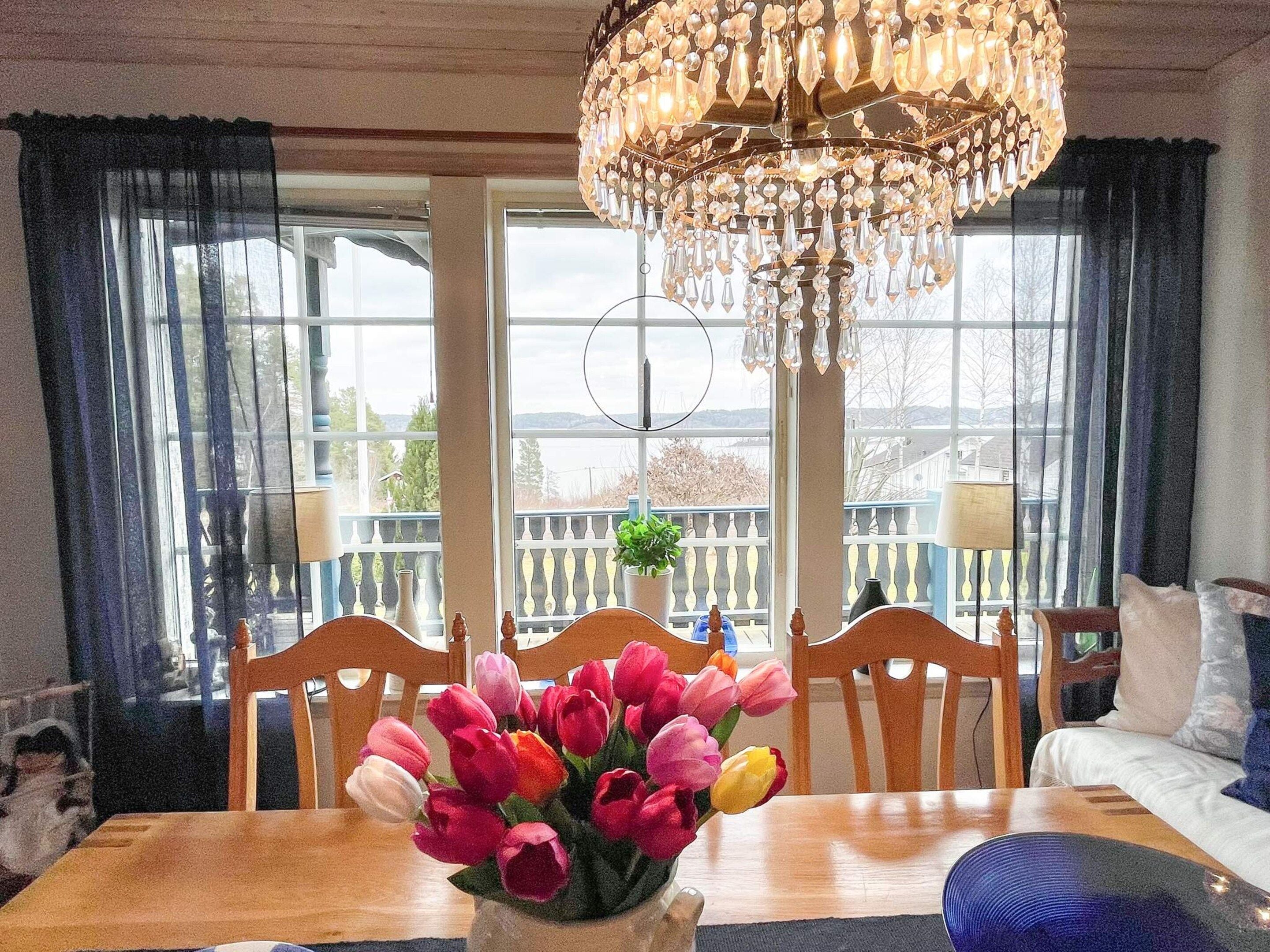 A dining area with a wooden table, chairs, a vase of tulips, a chandelier, and a view through large windows.