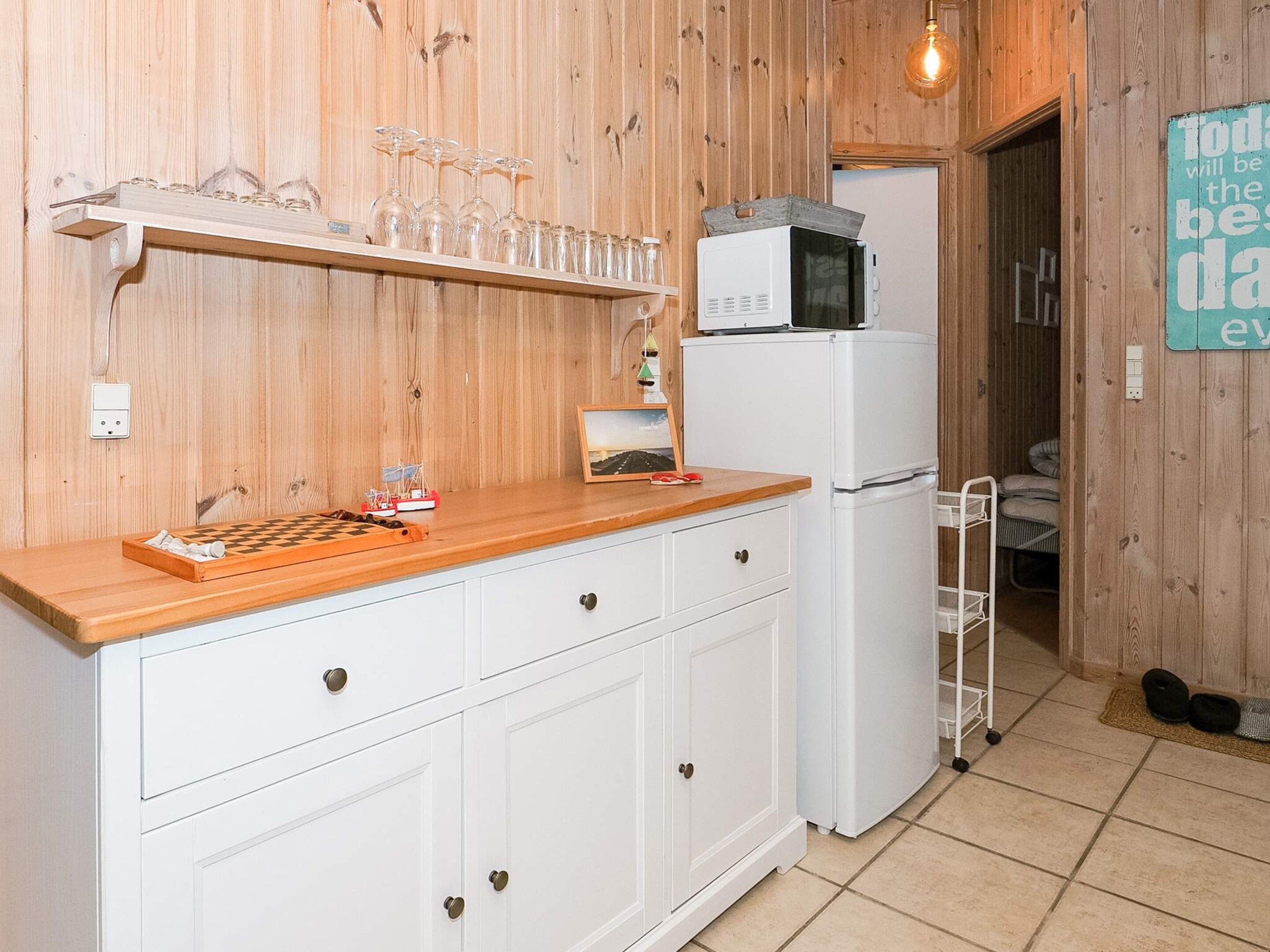 A kitchen with wooden walls, a white refrigerator, a microwave, a wooden countertop, a chessboard, and a shelf with glasses.