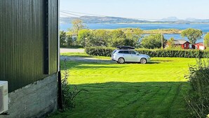 A view of a grassy field with a car parked in the distance, a body of water, and a red building in the background. - 6 Person Holiday Home in Vevelstad (Vevelstad)