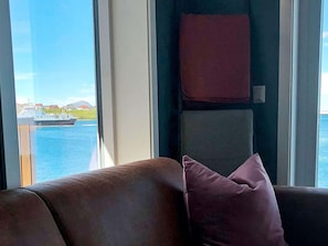 A room with a view of a body of water and a boat, a brown sofa, and a pink pillow.