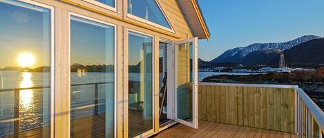 A wooden deck with glass doors leading to a balcony overlooking a body of water and a mountainous landscape.