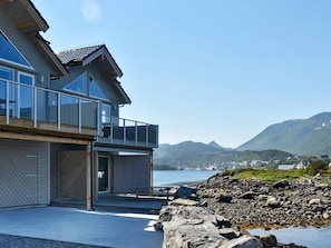 A modern house with a balcony overlooking a rocky shoreline and a body of water.
